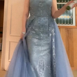 Ljusblå långklänning med spets och glitter - Säljer en magisk ljusblå/grå långklänning med glittriga detaljer och vacker spets. Klänningen har en elegant, figurnära passform upptill och ett svepande, voluminöst kjolparti i tyll. Perfekt för bal eller fest. Dragkedja i ryggen och fina broderade mönster framtill.