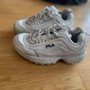Vita sneakers från Fila - Säljer ett par chunky vita sneakers från Fila med klassisk logga på sidan och plös. Skorna har snörning och en rejäl platt sula som ger extra höjd. Perfekta för dig som gillar sportig och retro stil.