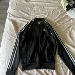 Svart Adidas track jacket med vita ränder - Klassisk svart Adidas track jacket med vita ränder längs ärmarna och vit Adidas-logga på bröstet. Jackan har dragkedja framtill, ribbade muddar och hög krage. Perfekt för en sportig och stilren look.