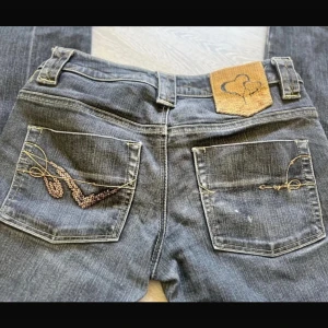 Mörkgrå jeansbyxor med paljettbroderi - Snygga mörkgrå jeans med dekorativa paljettdetaljer och broderi på bakfickorna. Byxorna har normal passform, klassisk femficksdesign och ett unikt märke i mockaimitation bak i midjan. Perfekta för dig som vill sticka ut med detaljer.