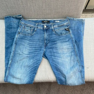 Blå Replay jeansbyxor - Säljer ett par klassiska blå jeans från Replay med fem fickor och snygga slitningar. Byxorna har raka ben och tydliga kontrastsömmar. Märkeslogga på bakfickan och läderpatch i midjan. Perfekta för dig som gillar en tidlös jeanslook.