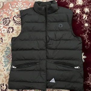 Svart dunväst från Moncler - Säljer en svart dunväst från Moncler med hög krage och två dragkedjefickor framtill. Västen har en quiltad design och Moncler-logga på bröstet. Perfekt för lager-på-lager under kyliga dagar. Medföljs tags samt påse! Storlek 2 motsvarar M