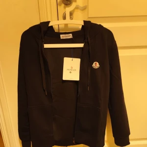 Moncler zip hoodie - Säljer en stilren svart hoodie från Moncler med dragkedja och huva. Perfekt för en avslappnad look. Den är storlek M men är liten i storleken så jag skulle själv kalla det för S.