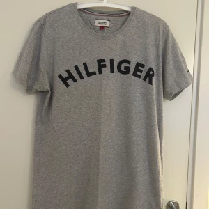 Grå t-shirt från Tommy Hilfiger - Grå t-shirt med svart tryck över bröstet från Tommy Hilfiger i storlek L. Rund halsringning och korta ärmar med en liten logga på ena ärmen. Adrig använd.