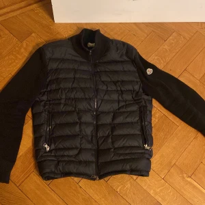 Svart pufferjacka från Moncler - Säljer en svart pufferjacka från Moncler med stickade ärmar och quiltad front. Jackan har dragkedja framtill och två fickor med dragkedja. Klassisk Moncler-logga på ena ärmen. 