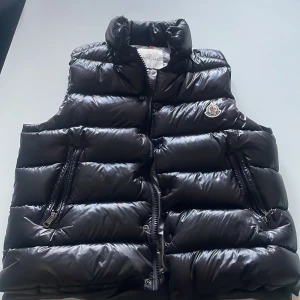 Svart dunväst från Moncler - Säljer en svart, glansig dunväst från Moncler med hög krage och två dragkedjefickor framtill. Västen har tryckknappar och dragkedja samt Moncler-logga på bröstet. Insidan har en serietidningsdetalj och klassisk Moncler-etikett. Och den är äkta.