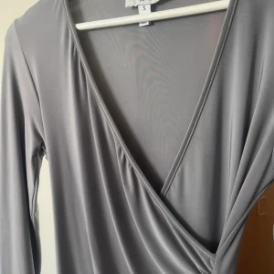 Grå omlottklänning med lång ärm från Rebecca Stella - Säljer en stilren grå omlottklänning med lång ärm och v-ringning. Klänningen har snygga draperingar framtill som ger en cool look. Perfekt för dig som gillar en enkel men trendig stil.