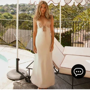 Champagnefärgad långklänning med knyt och v-ringning - Säljer en elegant champagnefärgad långklänning med smala axelband, v-ringning och knyt framtill samt i ryggen. Klänningen har en mjuk, flytande siluett och passar perfekt till festliga tillfällen.