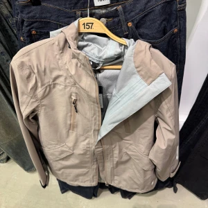 Vindjacka - Lätt och stilren beige vindjacka med huva och dragkedja framtill. Jackan har flera fickor med dragkedja och diskreta reflexdetaljer. Perfekt för blåsiga dagar och enkel att matcha med olika outfits.