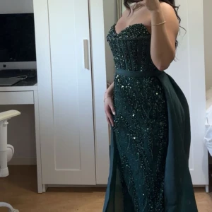 Mörkgrön glittrig långklänning med pärlor - Säljer en mörkgrön långklänning med glittriga detaljer och pärlor. Klänningen är axelbandslös och har en markerad midja samt en lätt utsvängd kjol. Den har en överkjol som medföljer, den är även avtagbar. Storleken är liten 36, Turkiet storlek som är lite mindre än vanligt. 