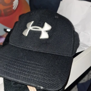Svart keps från Under Armour - Svart keps från Under Armour med stor vit broderad logga framtill. Klassisk böjd skärm och justerbar passform. Perfekt för sportiga tillfällen eller som en snygg accessoar.
