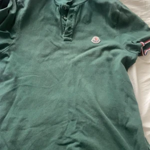 Grön Moncler pikétröja - Säljer en grön pikétröja från Moncler med klassisk krage och knappslå framtill. Tröjan har Moncler-logga broderad på bröstet och korta ärmar med en liten detalj i rött och vitt på ärmslutet. Perfekt för en stilren och sportig look.