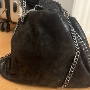 Svart axelväska med kedjedetaljer - Säljer en svart axelväska med glansig yta och kraftiga silverfärgade kedjor som axelrem. Väskan har en mjuk, säckliknande form och känns riktigt trendig och cool. Perfekt för dig som vill ha en statement-väska till din outfit.