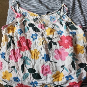 Blommig linnetopp med knappar - Söt linnetopp med färgglada blommor i rosa, blått och gult på vit botten. Toppen har smala axelband och små tygklädda knappar framtill. Perfekt för varma dagar och ger en somrig vibe.