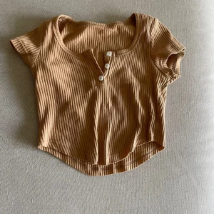 Beige ribbad croppad topp med knappar - Säljer en beige ribbad croppad topp med korta ärmar och rund halsringning. Toppen har tre dekorativa knappar framtill och är tillverkad i ett mjukt, stretchigt material. Perfekt för varma dagar eller som ett lagerplagg.