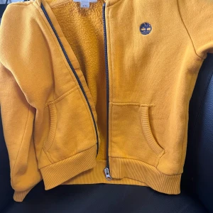 Timberland kofta (barn) - Säljer en gul hoodiejacka från Timberland med dragkedja och två fickor framtill. Timberland-logga broderad på bröstet. Perfekt för barn som gillar färg och vill ha något bekvämt och snyggt.