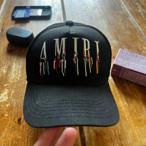 Svart keps från Amiri med broderad logga - Snygg svart keps från Amiri med broderad logga framtill i vitt och färgglada detaljer. Kepsen har böjd skärm, mesh baktill och justerbar snapback. Perfekt för en trendig streetwear-look.