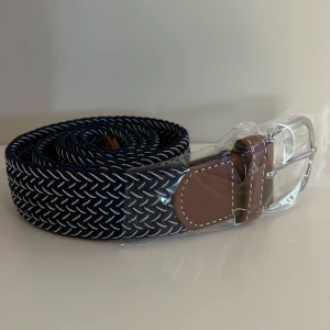 Blått/Vitt flätat bälte med bruna skinn-detaljer - Snyggt blått flätat bälte med silverfärgat spänne och bruna skinn-detaljer vid spännet. Bältet har ett diskret mönster i vitt och blått som ger en modern look. Perfekt accessoar för att lyfta din outfit.