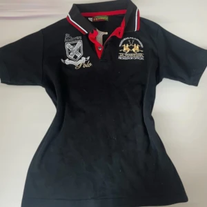 Svart pikétröja från La Martina - Svart pikétröja från La Martina med broderade detaljer och loggor på bröstet och ryggen. Kragen har röd och vit rand, och tröjan har korta ärmar samt knappslå framtill. Perfekt för dig som gillar sportig och stilren look.