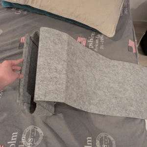 Grå filtad förvaring att hänga på sängen eller rliknande - Minimalistisk förvaringsutrymme i ljusgrå filt med enkel design och rektangulär form. Perfekt för att bära laptop eller dokument, med ett stort huvudfack och mjukt material som skyddar innehållet. Stilren och praktisk för att lägga bort skolmaterial i eller olika teknik prylar.