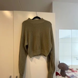 Olivgrön croppad stickad tröja - Säljer en olivgrön croppad stickad tröja med hög krage. Tröjan har lång ärm och en avslappnad passform, perfekt att styla med höga jeans eller kjol. Enkel och stilren design som passar till många tillfällen.