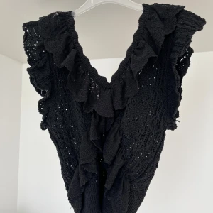 Svart volangtopp med spetsdetaljer - Säljer en svart topp med djup v-ringning och volanger längs halsringningen. Toppen har spetsdetaljer och är ärmlös, perfekt för att lyfta vilken outfit som helst.