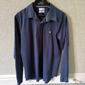 Mörkblå skjorta från Lacoste - Säljer en mörkblå långärmad skjorta från Lacoste i slim fit-modell. Skjortan har klassisk krage, knappar framtill och den ikoniska Lacoste-loggan broderad på bröstet. Perfekt för dig som gillar stilrena plagg med snygg passform. Verkligen perfekt för sommaren🔥