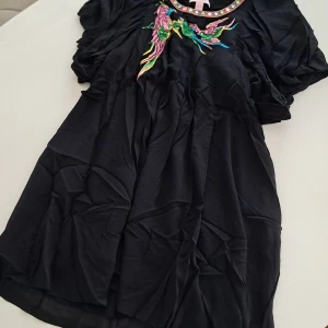 Svart silkesklänning Matthew Williamson H&M - Unik svart kortklänning i 100% siden från Matthew Williamson for H&M. Puffiga korta ärmar, färgglad broderad fågeldekor och coola detaljer runt halsen med pastellfärgade cirklar. Perfekt för dig som vill sticka ut med en statement piece!