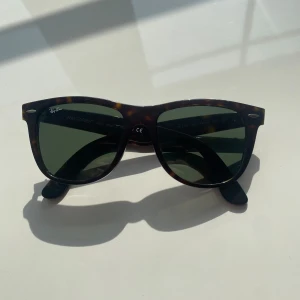 Ray-Ban Wayfarer brunmelerade solglasögon - Klassiska Ray-Ban Wayfarer solglasögon med brunmelerad båge och gröna glas. Snygg retrostil med ikonisk form och diskret logga på linsen. Perfekta för dig som vill ha en tidlös look med lite edge.