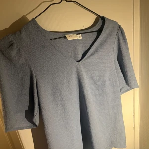 Ljusblå blus med v-ringning från Vila - Supersöt ljusblå blus från Vila med v-ringning och korta ärmar. Perfekt att styla med jeans eller kjol🤗 