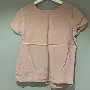 Ljusrosa blus i silkeslook - En ljusrosa blus med korta ärmar och rund halsringning framtill samt v-ringning med slits bak. Toppens material har en silkeslen känsla och lätt glans. Passar perfekt till jeans eller kjol för en stilren look.