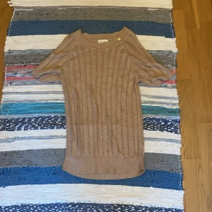 Beige stickad tröja med knappar - Snygg beige stickad tröja med korta ärmar och dekorativa knappar vid axeln. Tröjan har ett ribbat mönster och är perfekt för dig som gillar en enkel men trendig stil. Passar bra till både jeans och kjol.