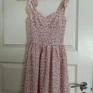 Blommig kortklänning från SHEIN XS - Supersöt kortklänning från SHEIN i storlek XS. Klänningen är ljusrosa med små blommor i rött och rosa, har breda axelband och markerad midja. Tillverkad i ett lätt bomullsliknande material och stängs med dragkedja bak. Perfekt för sommarens alla tillfällen!