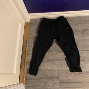 Svarta mjukisbyxor från Nike - Svarta mjukisbyxor från Nike med elastisk midja och dragsko. Byxorna har ribbade muddar vid bensluten och en diskret Nike-logga på benet. Perfekta för chill eller träning, tillverkade i mjukt bomullsmaterial för extra komfort.