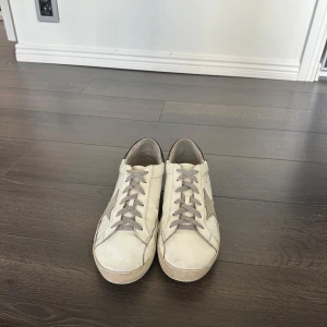 Golden Goose vita sneakers med stjärna - Golden Goose sneakers i vitt skinn med grå stjärna på sidan och grå snörning. Hälpartiet är svart med logga och sulan är platt. Klassisk låg modell med rund tå och cool vintagekänsla. Perfekta för dig som gillar streetstyle och snygga detaljer. Obs! Inget org som låda eller dustbag kommer med skorna!!