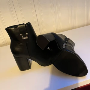 Svarta boots med klack och dragkedja - Snygga svarta boots i skinn med chunky klack och dragkedja på sidan. Skorna har en stilren design med rund tå och en metall-detalj upptill. Perfekta för dig som vill ha en klassisk men trendig look.