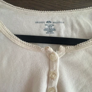 Vit topp med knappar från Brandy Melville - Supersöt vit topp från Brandy Melville i mjuk bomull. Rund halsringning med spetskant och tre knappar framtill ger en gullig och enkel vibe. Perfekt till jeans eller kjol för en chill look.