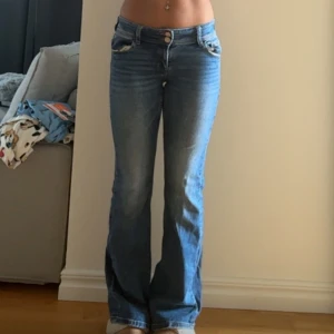 Lågmidjade blå bootcut jeans - Snygga lågmidjade jeans i mellanblå tvätt med bootcut-ben. Klassisk femficksmodell med ficklock och knapp bak. Jeansen har en lätt utsvängd passform och sitter tajt upptill. Perfekta för en avslappnad och trendig look.