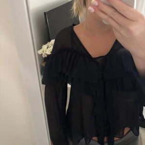 Svart volangblus i transparent material - Säljer en svart blus med volangdetaljer över axlar och bröst. Blusen är transparent och har långa ärmar med volang vid ärmslut. Perfekt för dig som gillar en luftig och trendig look. Ingen synlig logga eller märke.