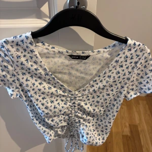 Blommig croppad topp från SHEIN - Supersöt croppad topp från SHEIN i vitt med små blå blommor. Toppen har korta ärmar, v-ringning och snörning framtill som ger en snygg rynkad effekt. Ribbad struktur i tyget och perfekt för vår och sommar.