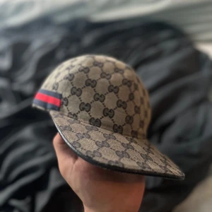 Beige keps från Gucci med GG-mönster - Snygg beige keps från Gucci med klassiskt GG-mönster över hela kepsen och en röd-blå rand på sidan. Justerbar rem baktill för perfekt passform. Perfekt accessoar för att lyfta din stil.