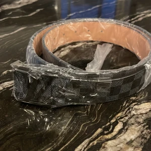 Svart och grått rutigt bälte - Snyggt bälte med svart och grått rutmönster. Bältet har en klassisk design med silverfärgat spänne och insida i beige. Perfekt accessoar för att lyfta din outfit.