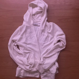 Vit hoodie från Wow - Säljer en vit hoodie från Wow med broderad logga på bröstet. Tröjan har huva, långa ärmar och fickor framtill. Perfekt för en avslappnad stil och enkel att matcha med det mesta.