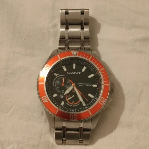 GANT herrklocka med orange bezel - Stilren herrklocka från GANT med robust länkarmband i silverfärgad metall. Urtavlan är svart med vita och orange detaljer samt en tydlig orange bezel. Klockan har kronograf och vattentäthet upp till 100M. Behövs byta batteri. 