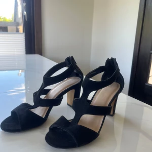 Svarta klackar med remmar från Shoes by din sko - Säljer ett par svarta sandaler med klack och flera breda remmar över foten. Skorna har öppen tå och dragkedja bak för enkel påtagning. De är i storlek 38 och användes 1 gång, så de är i princip som nya.