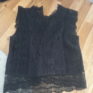 Svart spetsblus med v-ringning - Säljer en svart blus i spets med v-ringning och fina spetsdetaljer längs kanterna. Blusen är ärmlös och har ett vackert mönster samt dekorativ nederkant. Perfekt att styla till olika looks!