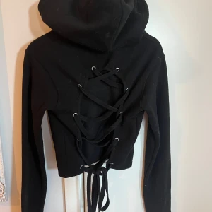 Svart croppad hoodie med snörning från Madlady - Säljer en svart croppad hoodie från Madlady med långa ärmar och snygg snörning i ryggen. Perfekt för dig som vill ha en unik och edgy stil. Mjuk och skön, passar till många outfits.