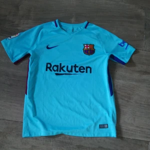 FC Barcelona ljusblå bortatröja Nike - Snygg ljusblå FC Barcelona bortatröja från Nike med klubbmärke och Rakuten-tryck på bröstet. Kortärmad modell med rund hals och detaljer i mörkblått och vinrött på ärmarna. Materialet är lätt och andas, perfekt för fotbollsträning eller match.