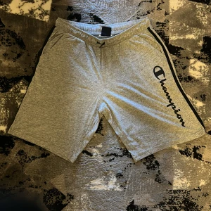 Grå Champion shorts med snörning - Grå mjuka shorts från Champion med svart logga längs ena benet och svarta detaljer på sidorna. Elastisk midja med snörning för skön passform. Perfekta för träning eller chill, tillverkade i bomullsmix. Klassisk och sportig look.