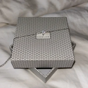 Silverfärgat armband från Edblad - Säljer ett stilrent silverfärgat armband från Edblad med tunn kedja och en liten rund bricka med logotyp samt en gnistrande sten. Perfekt för dig som gillar minimalistiska smycken.